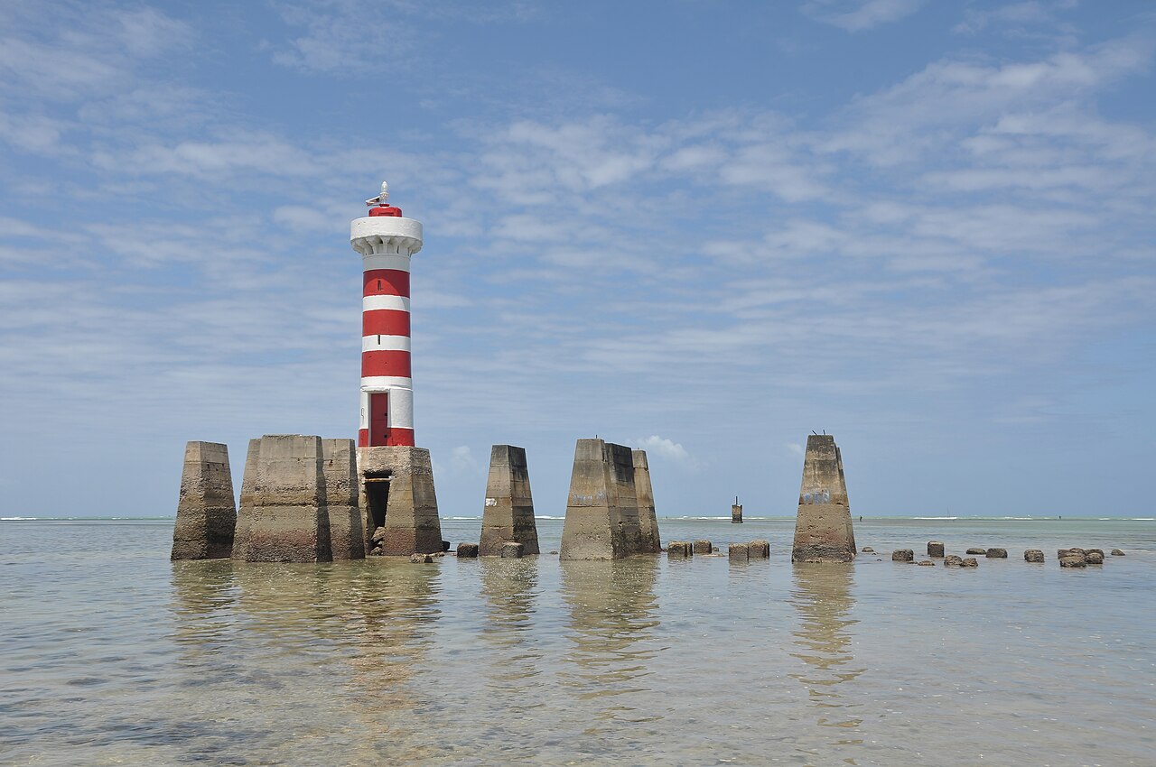 Farol da Ponta Verde em águas rasas, com barcos ao fundo, em Maceió